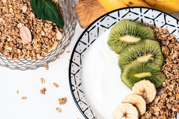 Overhead view of a breakfast bowl with yogurt, cereals and sliced ​​kiwi pieces with a wooden board with bananas and another glass bowl with cereals around

