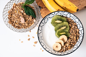Breakfast bowl with yogurt, cereal and sliced pieces of kiwi and banana with another glass bowl next to it full of cereal and two bananas on a wooden board
