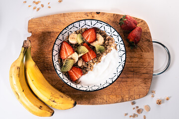 Overhead view of a breakfast bowl filled with yogurt, cereal and sliced pieces of strawberries, banana and kiwi on a wooden board with two bananas and two strawberries around
