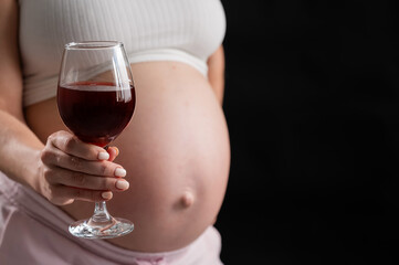 Close-up of the belly of a pregnant woman holding a glass of red wine on a black background. 