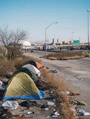The image captures a roadside encampment with several tents. There is a significant amount of litter and debris scattered around the area. In the background, there is a highway with traffic signs. - obrazy, fototapety, plakaty