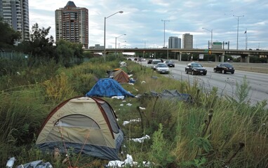 A sprawling makeshift encampment has taken root along the highway, surrounded by the imposing high-rise buildings of the urban skyline, highlighting the stark divide between affluence and homelessness - obrazy, fototapety, plakaty
