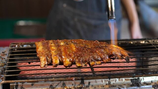 The chef is grilling pork ribs with BBQ sauce on the stove and using a flame to burn the ribs.