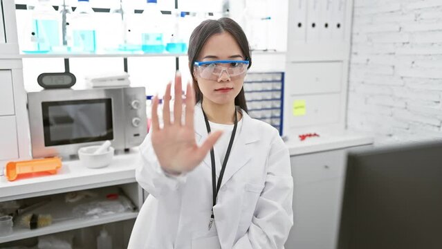 Confident young chinese scientist woman inside lab, rejecting with serious no-go warning gesture using her palm. expresses negative face reaction