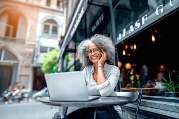 Cheerful woman sits in a coffee shop with her laptop and mobile phone, multitasking and getting work done in a relaxed setting. Young female freelancer working online.