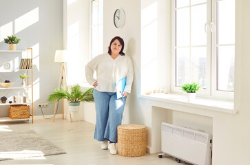 Young overweight woman in casual clothes standing leaning wall. Attractive plump, plus size brunette woman holding paper documents posing at home or office and smiling at camera