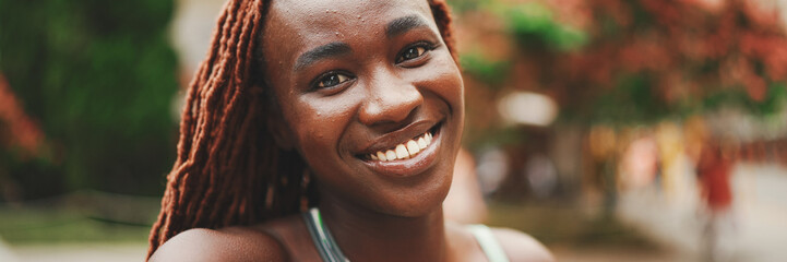Clouse-up, smiling gorgeous woman with African braids wearing top stands outside on the street and...