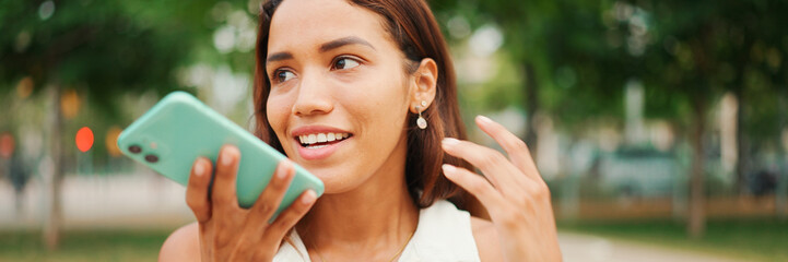 Beautiful girl with long dark hair wearing white top sits on bench and uses mobile phone, Panorama....