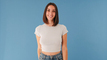 Beautiful brunette young woman dressed in white top looking at camera and smiling widely isolated on blue background in studio