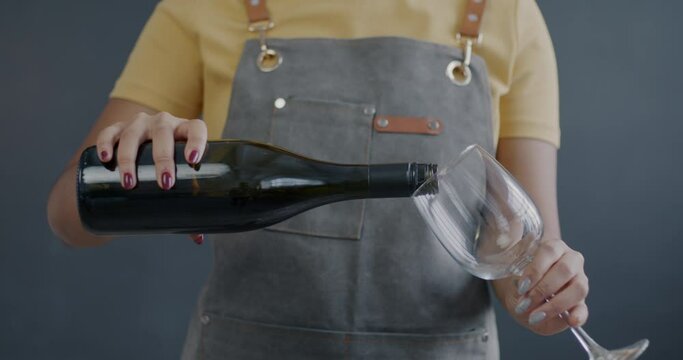 Close-up of female hands pouring red wine from bottle in glass while waitress serving alcohol on grey color background. Winery business and careting concept.