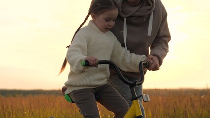 Woman teaching girl to ride bike, silhouettes at sunrise in field. Emotional support, participation, presence and encouragement from mother in learning process to help child overcome fear of failure. - obrazy, fototapety, plakaty
