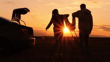 Silhouettes of having fun mom dad kid. Golden sunset, cheerful joyful family at holidays holding hands, lifting baby girl high in air, playing together. Fun active game. Traveling by car to nature. - obrazy, fototapety, plakaty