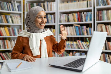 A cheerful Muslim woman wearing a hijab uses a laptop in a library setting, waving and smiling,...