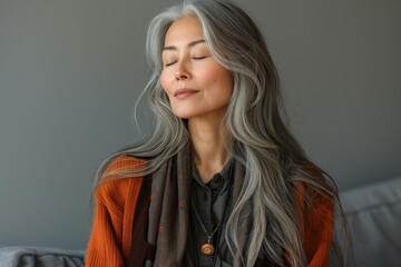 A mature Asian woman looks peaceful with eyes closed, wearing scarf and sweater in a comfortable setting - obrazy, fototapety, plakaty