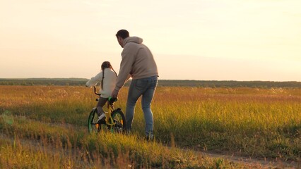 Parent teaches child to ride bicycle. Father and daughter in field at epic sunset. Child safety, dad regulates and controls beloved girl kid on bike, warning against injuries and bruises. Slow motion - obrazy, fototapety, plakaty