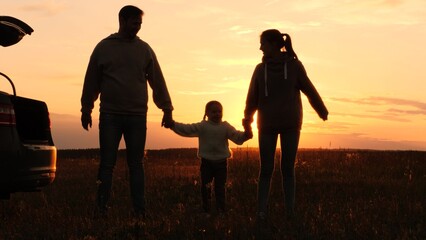 Family holding hands at sunset, silhouettes near car. Joint recreation in nature. Mother father daughter in orange setting sun in field. Family picnic road trip, parents kid enjoy each other's company - obrazy, fototapety, plakaty