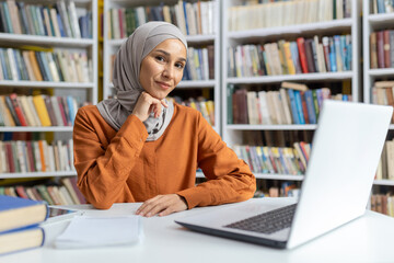 A poised Muslim woman with a hijab sits at a desk in a library, engaging with a laptop, surrounded by books. She appears focused and professional.