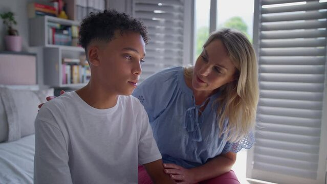 Mother talking with and comforting unhappy teenage son with problem sitting on bed at home - shot in slow motion