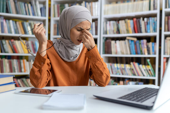 A young woman in a hijab sits at a library desk, feeling stressed and touching her temples, surrounded by books and a laptop.