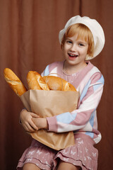 Girl in colorful outfit and white hat, holding a paper bag filled with various types of bread....