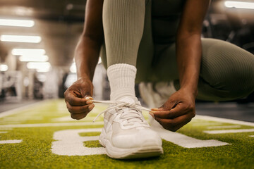 Close up of african american sportswoman's hands tying shoelace on sneaker at gym. - obrazy, fototapety, plakaty