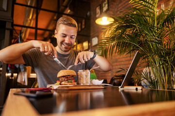 Smiling hungry man cutting and eating burger at restaurant.