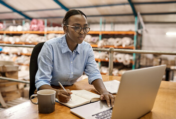 African business woman sitting at her warehouse desk working on a laptop