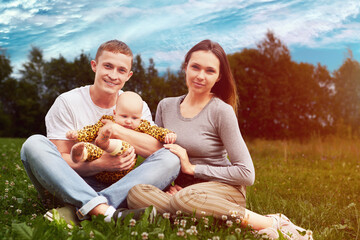 Twenty year old married couple with baby is sitting on grass of country lawn.