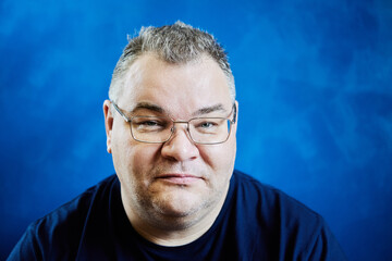 Friendly face of bespectacled white man in his 40s, overweight, close-up on blue background.