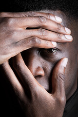A black man covers his face with his hand, revealing one eye and expressing contemplation.