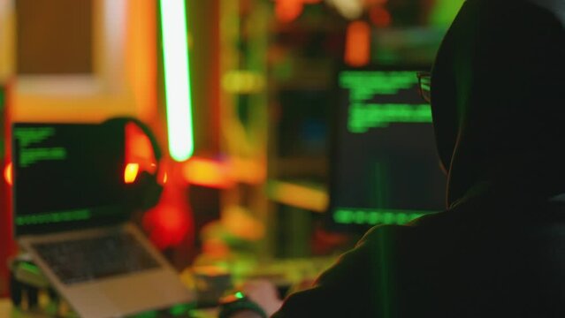 Hacker writing code cracking computer. Man programmer in hoodie cracks system, using computer in dark room back view. Hacker attack, technology, virus infected software, cyber security concept.