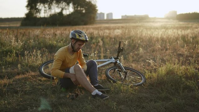 Sad white man bandaging knee after falling from bicycle. First aid for injured cyclist with helmet, suffering due to pain. Unsuccessful bike ride in picturesque field at sunset and fixation of joint.