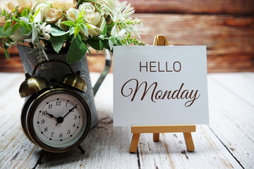 Hello Monday text message written on paper card with wooden easel and alarm clock with flower in metal vase decoration - obrazy, fototapety, plakaty