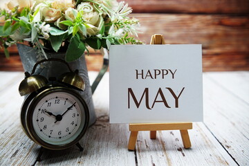 Happy May text message written on paper card with wooden easel and alarm clock with flower in metal...