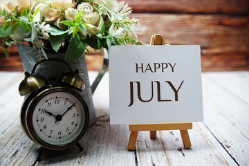 Happy July text message written on paper card with wooden easel and alarm clock with flower in metal vase decoration