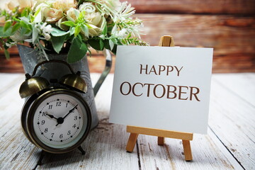 Happy October text message written on paper card with wooden easel and alarm clock with flower in...