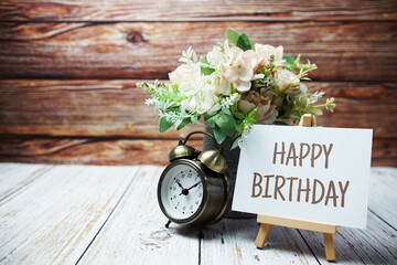Happy Birthday text message written on paper card with wooden easel and alarm clock with flower in...