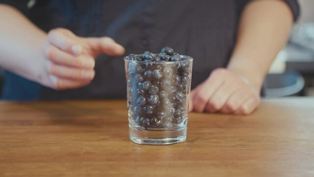 Woman picks up a glass of fresh blueberries. Close up of freshly washed blueberries in a glass on a wooden table in the kitchen. Defocused bright background. Vegan meal. High quality 4k footage