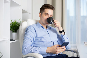 Young handsome business man sitting in his office, reading book and drinking tea
