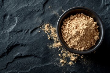 A bowl of maca powder on a textured black slate surface, with some powder sprinkled around
