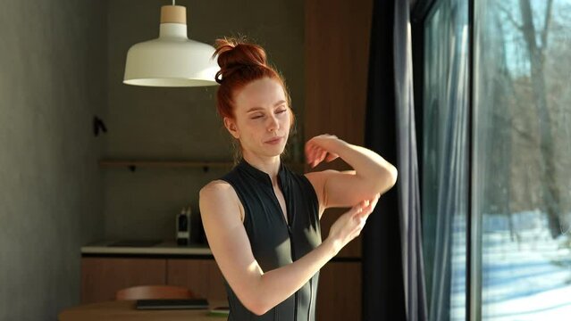 Focused athletic young woman in outfit preforming warming up exercise, stretching muscles before workout indoors, standing by window with snowy winter nature view on sunny day. Shooting in slow motion