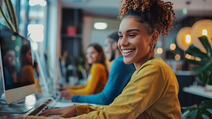 Smiling young woman with curly hair using computer in a vibrant coworking space. Casual business environment with colleagues