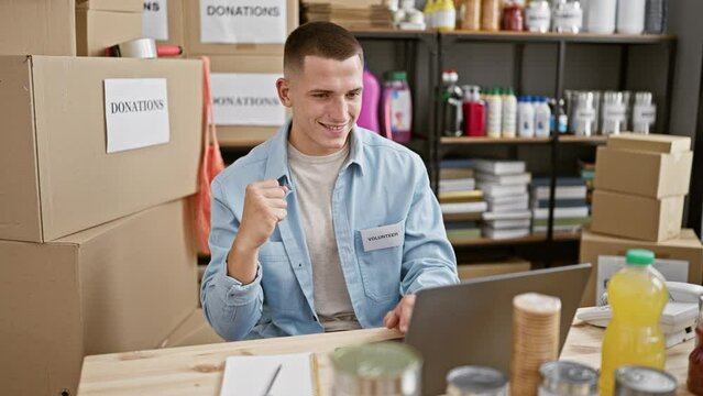 A young hispanic man volunteers at a donation center, organizing items while working on a laptop in a warehouse.