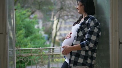 Happy pregnant woman gently caresses belly standing by window at apartment balcony overlooking view...