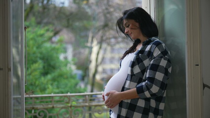 Happy pregnant woman gently caresses belly standing by window at apartment balcony overlooking view...