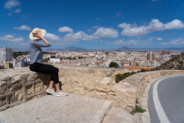 Woman in hat looking towards Alicante city. Castle of Santa Barbara area. Beautiful weather, blue...