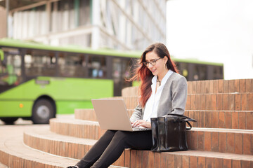 Latin young woman sitting outdoors in a city square on a sunny day with a laptop and a public bus in the background.