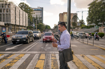 A young man crossing the street of a big city with a cell phone and a  coffee cup in his hands.