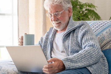 Senior bearded smiling man sitting on sofa looking at laptop holding a coffee cup - indoor, at home...