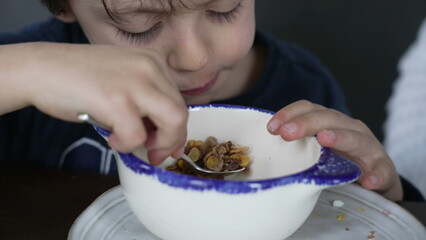 Small boy eating cereal with spoon inside bowl. Candid hungry child snacking wheat food for breakfast